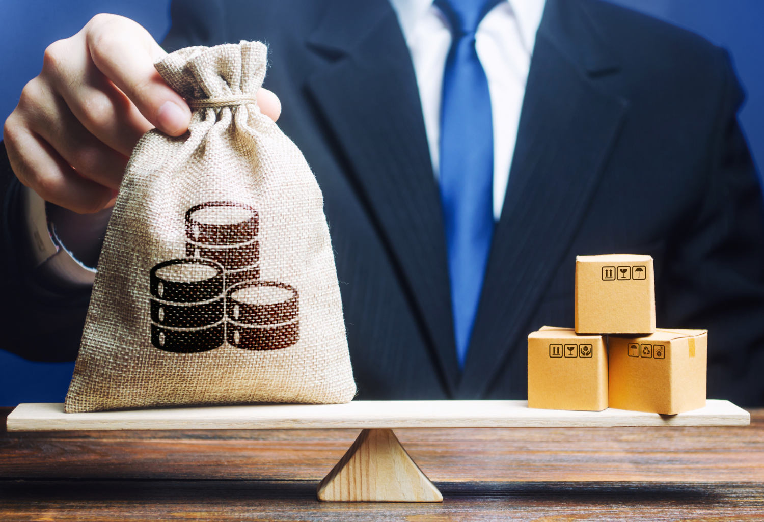 Suited man holding a coin bag on a see-saw with boxes on the other side